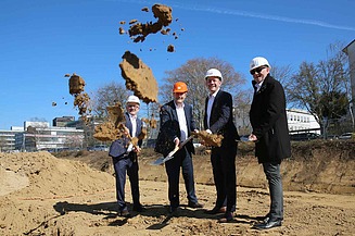 Foto von vier Männern in Anzug und einem Helm auf. Sie sind auf einer Baustelle und schaufeln Erde mit einem Spart.    