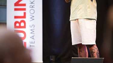 Foto von zwei Personen auf der Bühne. Sie spricht in ein Mikrophon.  