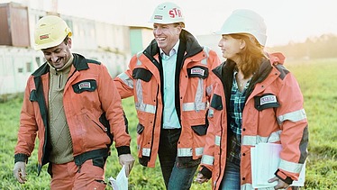 Foto von drei Personen in Arbeitskleidung auf einer Baustelle, die gut gelaunt wirken und Unterlagen bei sich tragen