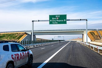 Das Bild zeigt ein weißes Auto mit der Aufschrift STRABAG auf einer Autobahn, im Hintergrund eine grüne Verkehrstafel und eine querende Brücke