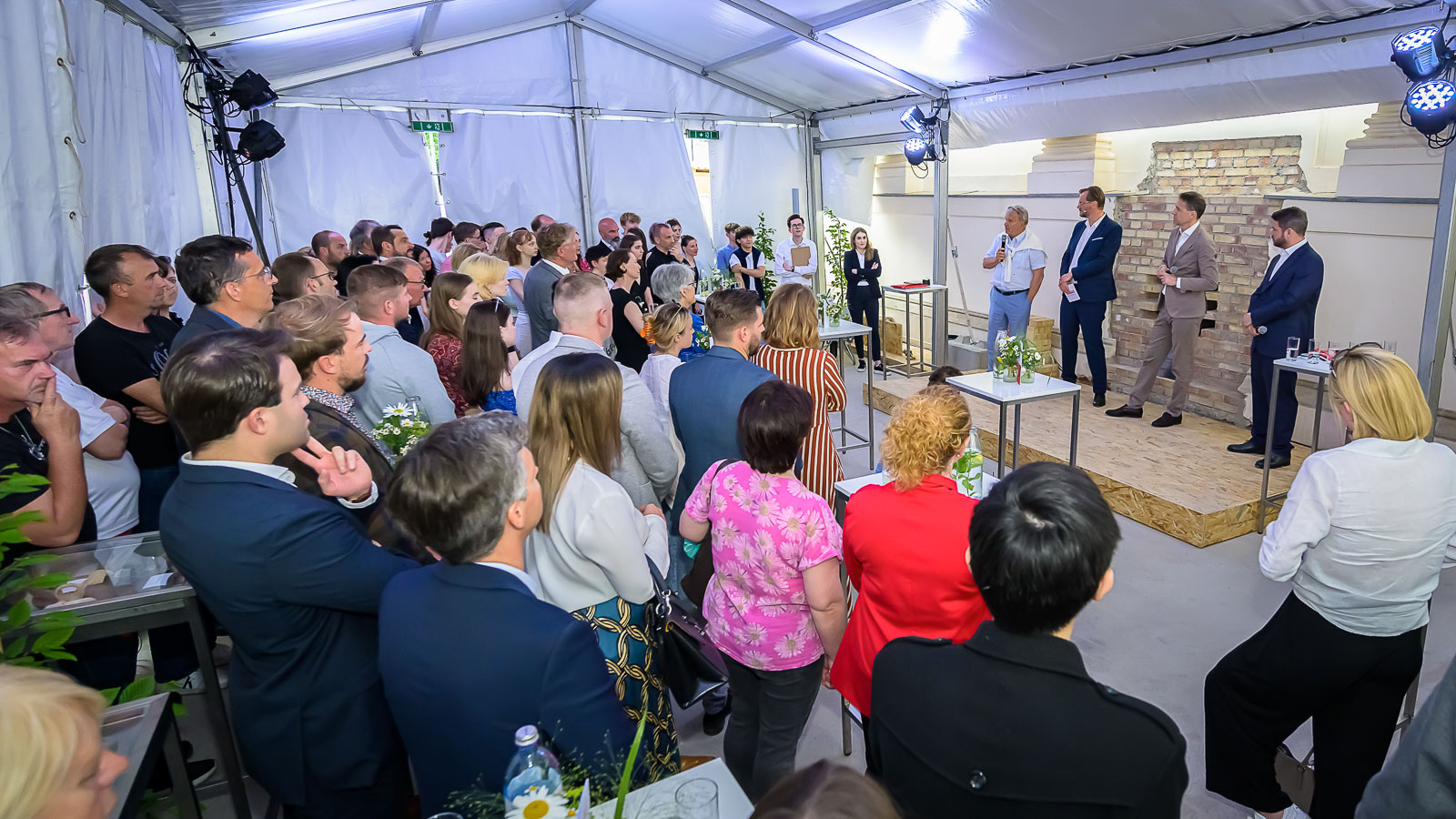 Foto von drei Personen auf einer Bühne bei der Grundsteinlegung des Franzoesischen Saals, davor stehen eine Ansammlung von vielen Personen