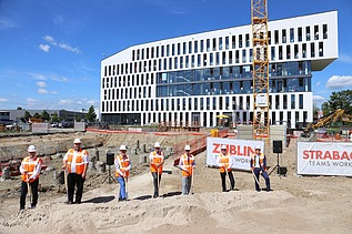 Foto von mehreren Männern in Warnweste auf einer Baustelle. Sie halten einen Spart in der Hand, der in den Boden gesteckt ist. Die Baustelle befindet sich vor einem Bürogebäude. 
