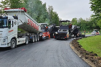 Foto von einem STRABAG LKW beim Abladen, daneben eine Asphaltbaumaschine