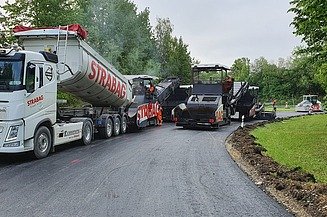 Foto von einem STRABAG LKW beim Abladen, daneben eine Asphaltbaumaschine