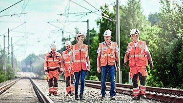 Foto von fünf Personen, die zwischen zwei Bahngleisen mit Oberleitungen gehen
