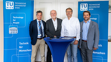 Foto von vier Personen, die an einem Stehtisch stehen und dahinter zwei blaue Roll-ups