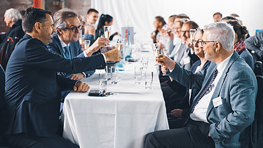 Foto von mehreren Personen bei der Jubiläumsfeier am Standort Sande. Sie sitzen an einem Tisch und stoßen an. 