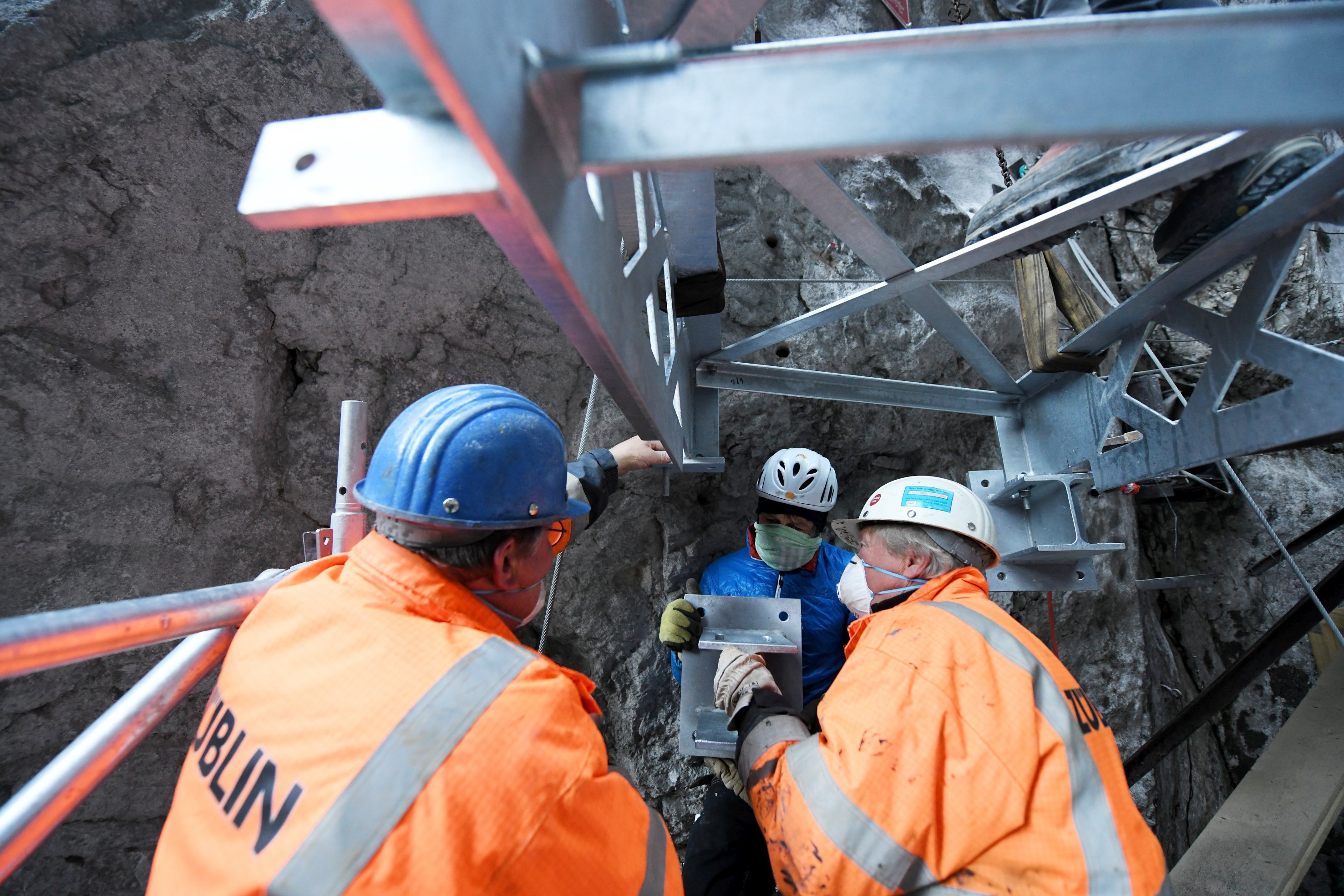 Foto von drei Personen in Schutzuniform und Mund-Nase Maske die an einem Stahlträger arbeiten.     