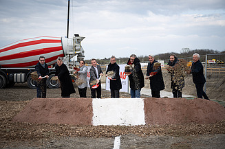 Foto von mehreren Personen beim symbolischen Spatenstich, links hinten steht ein LKW Betonmischer, der rot-weiß gestreift sit