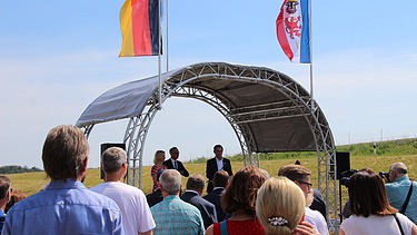 Foto von mehreren Personen, die vor einer überdachten Bühne stehen, dahinter zwei Flaggen