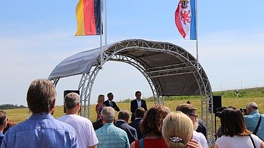 Foto von mehreren Personen, die vor einer überdachten Bühne stehen, dahinter zwei Flaggen