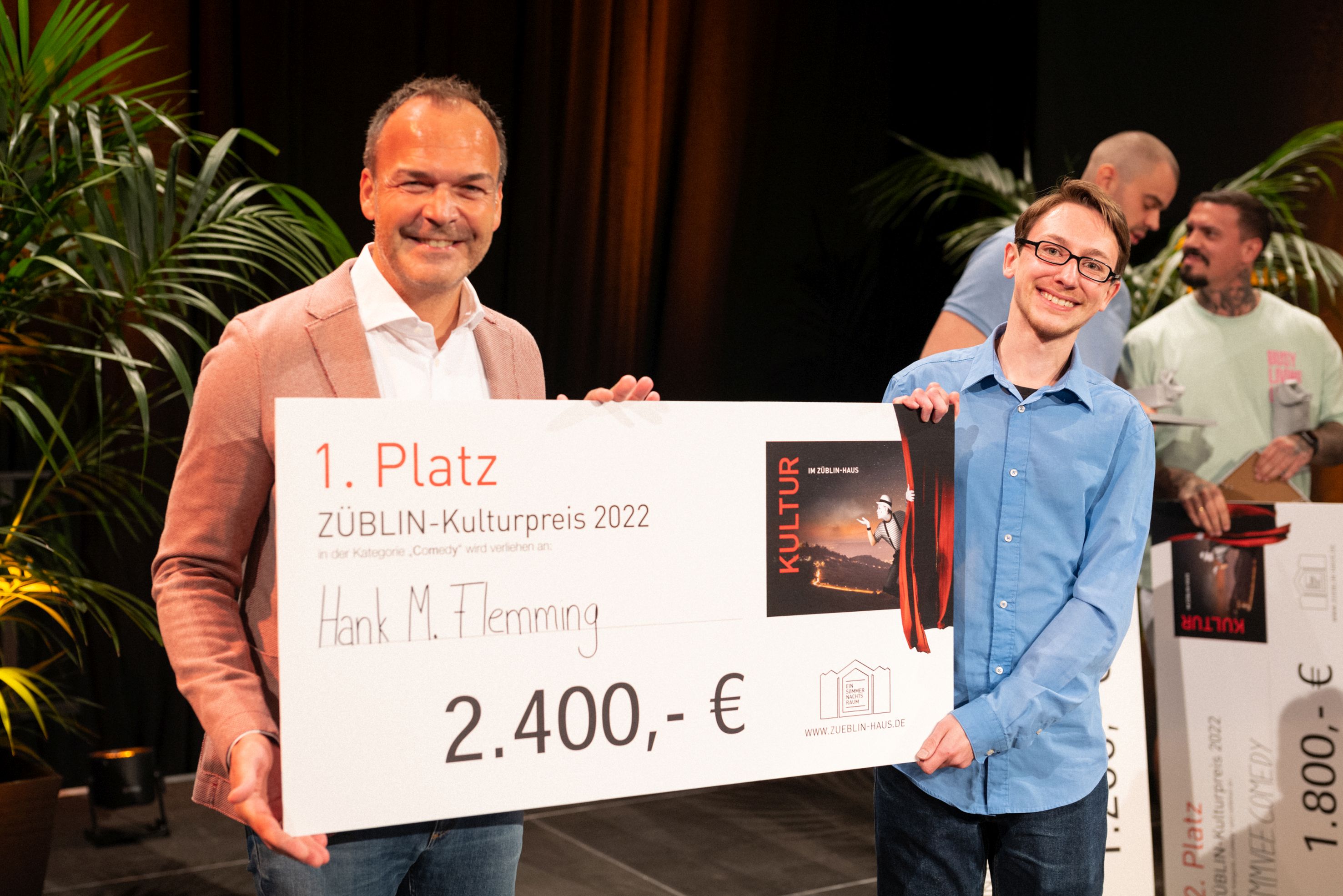 Foto von zwei Personen, die freundlich schauen. Sie halten den Preis zum ersten Platz des Comedy-Battles. 