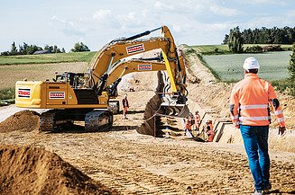 STRABAG-Mitarbeiter geht auf Bagger zu, der Leitungen verlegt