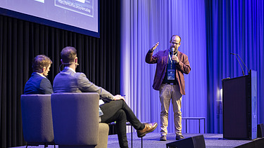 Foto von drei Personen auf einer Bühne, die Person rechts steht, die anderen Beiden sitzen in Couchsesseln