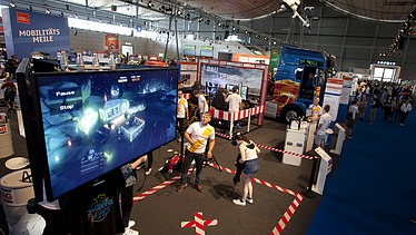 Foto von einem Großbildschirm in einer Messehalle, davor stehen zwei Personen