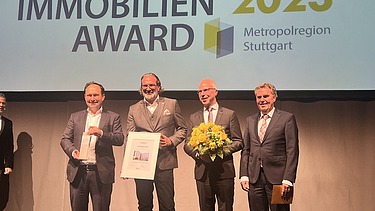 Foto von vier Männern im Anzug die auf einer Bühne stehen. 