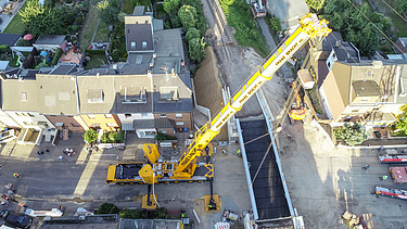 Foto von einem Wohngebiet, wo ein Kransteht der ein Teil für eine Brücke hält.  