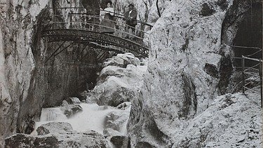 Schwarz-weiß Foto von einer Brücke, die über einen Graben geht. Drumherum sind Bergwände, die sich zu einer Schlucht formen. Auf der Brücke sind zwei Personen zu sehen. 
