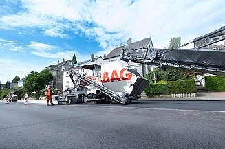 Foto einer Asphaltbaumaschine mit der Aufschrift STRABAG, sie steht am Straßenrand, der Himmel ist bewölkt