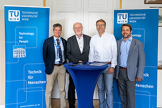 Foto von vier Personen, die an einem Stehtisch stehen und dahinter zwei blaue Roll-ups