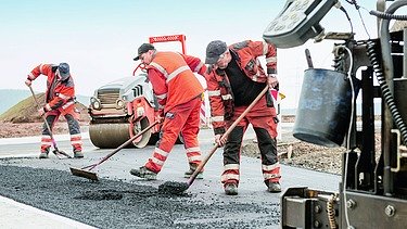 Foto von drei Straßenbauarbeitern mit Besen bei der Arbeit, rechts im Vordergrund sieht man einen Teil einer Straßenbaumaschine