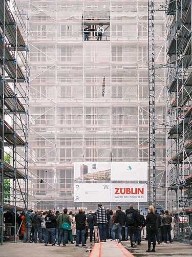 Foto von mehren Personen auf einer Baustelle. Auf einem Gerüst wird der Richtspruch verkündet. 