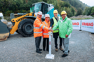 Photo of four people wearing construction helmets and protective jackets standing in front of a wheeled excavator