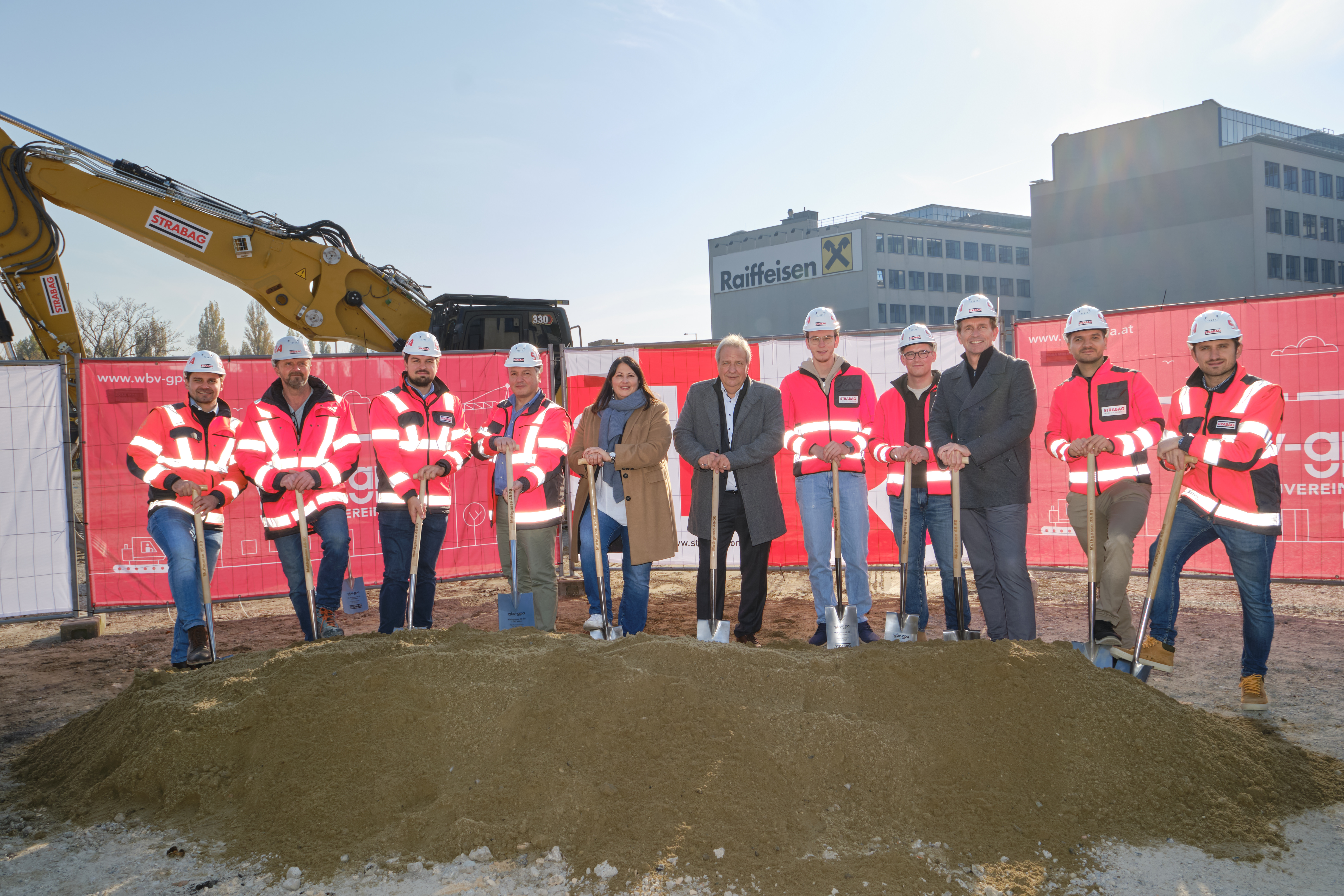 Photo of eleven people breaking ground, behind them you can see an excavator and two houses