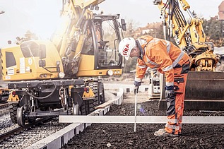 Foto von einem Gleisbauarbeiter in Schutzkleidung, dahinter eine gelbe Gleisbaumaschine