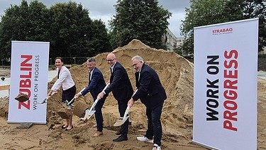 The picture shows four people with shovels at the ground-breaking ceremony, with two company roll-ups to the left and right