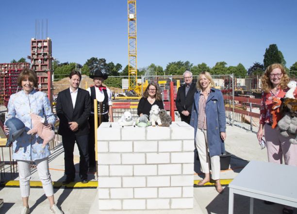 Foto von mehren Personen auf einer Baustelle, sie sind schick gekleidet und haben Kuscheltiere in der Hand. Sie stehen um eine kleine Mauer auf denen ebenfalls Kuscheltiere liegen.  