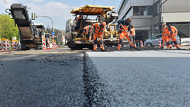 Foto einer frisch aspahltierten Straße, im Hintergrund sieht man zwei Straßenbaumaschinen