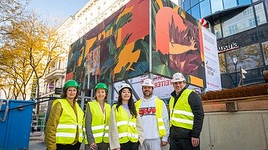 Foto von fünf Personen mit gelben Schutzwesten und Bauhelmen vor einer Baustelle, die mit einem Kunsttransparent umhüllt ist