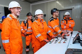 Photo of several people wearing hard hats and orange protective clothing standing at a control panel