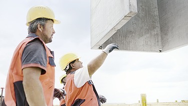 Foto von zwei Personen, die in Arbeitsuniform einen Betonblock navigieren, dass er nach oben gezogen wird. 