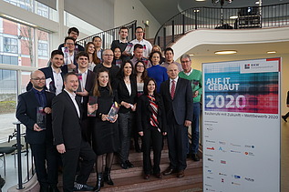 Foto von mehreren Personen auf einer Treppe. Sie sind schick angezogen und halten ein Pokal in den Händen. Neben ihnen ist ein Banner von Bauberufe mit Zukunft – Wettbewerb 2020.     