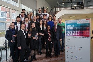 Foto von mehreren Personen auf einer Treppe. Sie sind schick angezogen und halten ein Pokal in den Händen. Neben ihnen ist ein Banner von Bauberufe mit Zukunft – Wettbewerb 2020.     