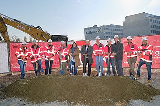 Photo of eleven people breaking ground, behind them you can see an excavator and two houses