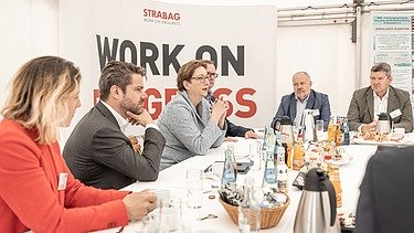 Foto von mehreren Personen, die an einem weißen Tisch sitzen, dahinter ein Transparent mit der Aufschrift Work on Progress