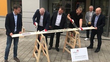 Foto von mehreren Männern im Anzug, die ein Brett sägen. 