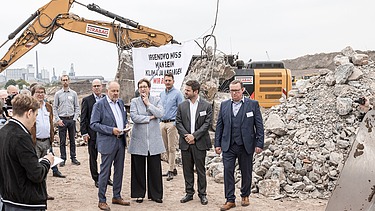 Photo of several people visiting a construction site, in the background a yellow excavator at work