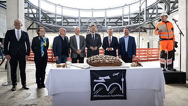 Sieben gute Gelaunte Personen hinter einem Tisch, der mit einem großem Brot gedeckt ist. Rechts von ihnen ein Mann der in Arbeitsuniform eine Rede hält.