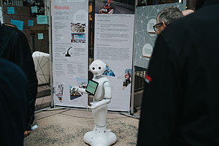 Photo of a small, white robot standing in front of a trade fair booth