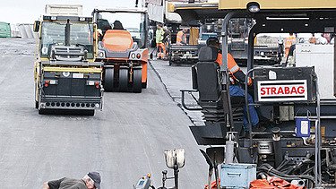 Foto von mehreren Straßenbaumaschinen und einem Arbeiter mit einer Schaufel