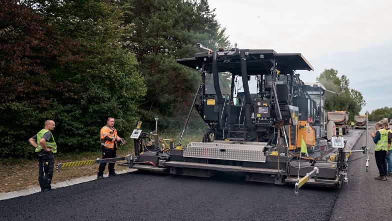 Foto von einer Asphalteinbaumaschine beim Einsatz, einige Personen stehen an beiden Seiten