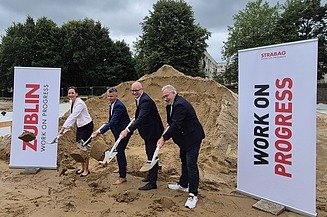The picture shows four people with shovels at the ground-breaking ceremony, with two company roll-ups to the left and right