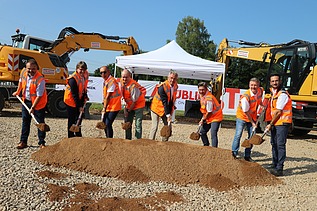 Foto von mehreren Personen bei sonnigem Wetter auf einer Baustelle, die jeweils einen Spaten in den Händen halten und einen symbolischen ‚Spatenstich‘ ausführen