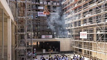 Foto von einer Baustelle, wo der Richtkranz aufgehangen wird. Viele Menschen schauen sich, dies an.      