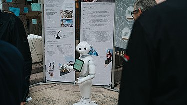 Photo of a small, white robot standing in front of a trade fair booth