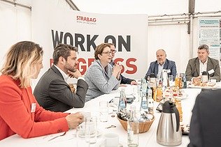 Foto von mehreren Personen, die an einem weißen Tisch sitzen, dahinter ein Transparent mit der Aufschrift Work on Progress